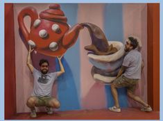 two men in front of a large painting with teapots on the wall and one man kneeling down