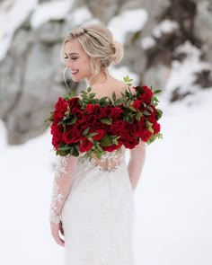 a woman in a wedding dress holding a bouquet of red roses and greenery on her shoulder