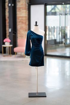 A mannequin in a cocktail dress showcases a fitted silhouette with ruched velvet fabric.  The classic one shoulder bishop sleeve with embellished jeweled cuff adds just the right amount of detail. Elegant One-shoulder Velvet Party Dress, Elegant Velvet One-shoulder Dress, Elegant One-shoulder Velvet Evening Dress, One-shoulder Velvet Cocktail Dress, One Shoulder Fitted Velvet Party Dress, Fitted One-shoulder Velvet Party Dress, Fitted One-shoulder Velvet Dress For Party, Elegant One-shoulder Velvet Dress, Formal Holiday Dress