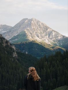 a woman standing on top of a lush green hillside next to a forest covered mountain