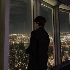 a man standing in front of a window looking out at the city lights and skyscrapers