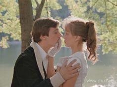 a man and woman are kissing in front of some trees by the water with their arms around each other