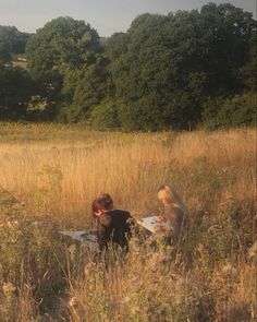 two people sitting on a blanket in the middle of a field with trees behind them