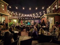 a group of people sitting at tables in front of a building with lights strung over it