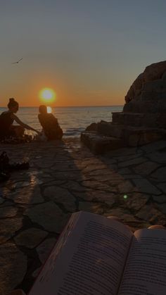 an open book sitting on top of a stone walkway next to the ocean at sunset