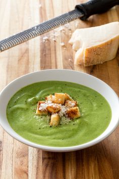 a white bowl filled with green soup on top of a wooden table next to bread