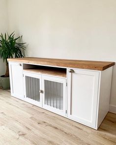 a white entertainment center with wooden top and glass doors on the bottom, next to a potted plant