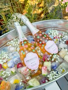 two bottles of alcohol sitting on top of ice cubes in a metal bowl filled with flowers