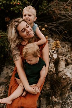 a woman holding a small child in her arms and smiling at the camera while standing on some rocks