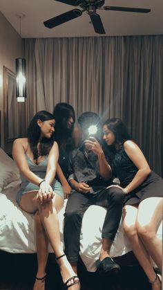 three women are sitting on a bed looking at a cell phone while the man is taking a selfie