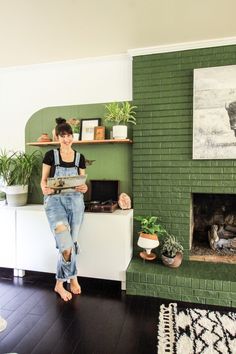 a woman sitting in front of a green brick fireplace with potted plants on the mantel