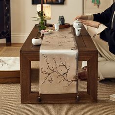 a woman is sitting at a table with a tea set on it and she is holding a remote control in her hand