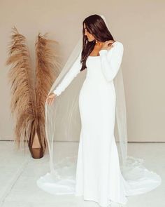 a woman wearing a white wedding gown with a long veil on her head standing in front of a tall plant