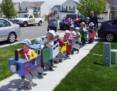 children are lined up in toy train cars on the sidewalk while adults watch from behind them