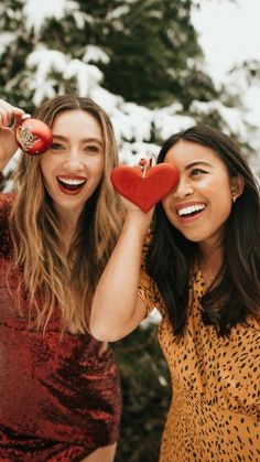 two beautiful women standing next to each other in front of snow covered trees and holding red hearts