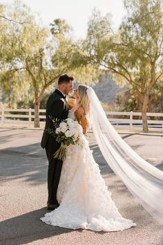 a bride and groom standing in front of a white fence with their veil blowing in the wind