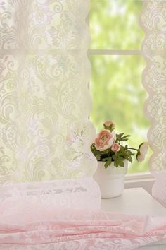 a white vase with pink flowers sitting on a window sill next to a curtain