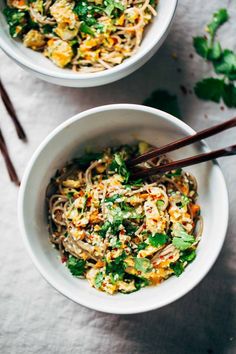 two bowls filled with noodles and vegetables on top of a white cloth next to chopsticks