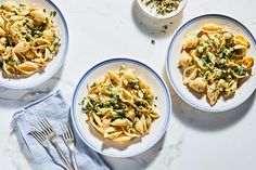 three white bowls filled with pasta on top of a marble table next to silverware