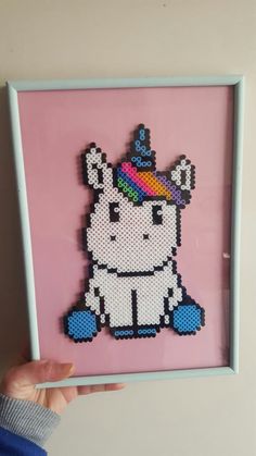 a person holding up a cross stitch picture in front of a pink wall with a unicorn on it