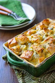 a green casserole dish filled with bread and vegetables on a wooden table next to two plates