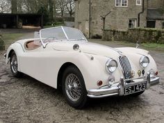 an old white sports car parked in front of a stone building with a clock on it's face