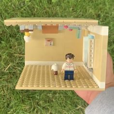 a person holding up a lego model of a house in the grass with a doll inside