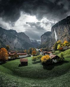 a green field with houses in the middle and mountains in the background under a cloudy sky