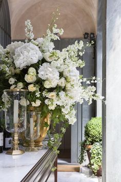 white flowers and greenery are on a table in front of an arched doorway with glass vases