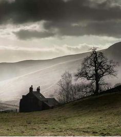 a house in the middle of a grassy field with mountains in the background and dark clouds overhead