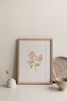 a pink flower is displayed in a wooden frame next to a white cup and saucer