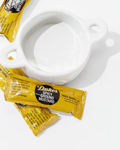 A high-fired ceramic dish, named Bowl No. 5 from Montes Doggett, sits on a white surface with three yellow packets of Duke's Spicy Ground Mustard placed beside and on top of it. The handmade dish, crafted in Peru, complements the sealed packets clearly labeled with ingredient information and branding. Plant Vessels, Pantry Furniture, Table Lamps Kitchen, Lamps Kitchen, Chandelier Table Lamp, Hand Molding, Ottoman Stool, Home Goods Decor, Candle Containers