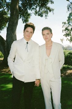 two young men in white suits standing next to each other under a tree on the grass