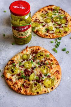 two small pizzas with pickles on them next to a jar of jalapenos