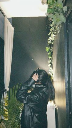 a woman in a black leather jacket is talking on her cell phone while standing next to a plant