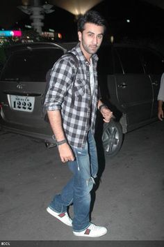 a man in plaid shirt and jeans walking on street next to parked car at night