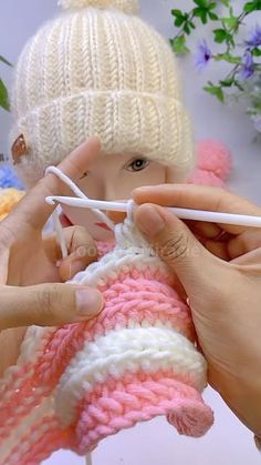 two hands holding knitting needles over a knitted baby's hat