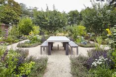 a table and bench in a garden with lots of flowers on the ground, surrounded by trees