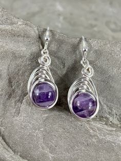 These beautiful genuine natural Amethyst earrings are truly unique. The violet purple color of the stone is beautiful in its simplicity. These amethysts smooth polished rounds that I chose to wire wrap in a simple herringbone design with silver plated wire to let the stone shine . These earrings are truly lovely. Each genuine Amethyst gemstone has been carefully selected for these earrings. They are made completely by hand by me. Pleasingly simple yet extraordinary, these earrings can be worn dr Handmade Amethyst Purple Earrings, Silver Amethyst Wire Wrapped Earrings, Round Purple Earrings With Natural Stones, Purple Natural Stones Round Earrings, Purple Earrings With Natural Stones, Lavender Earrings With Natural Stones For Gift, Lavender Amethyst Earrings With Natural Stones, Nickel-free Amethyst Earrings For Healing, Teardrop Purple Earrings With Natural Stones