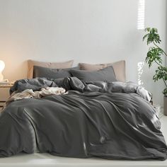 a bed with grey sheets and pillows in a room next to a plant on the floor