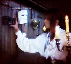 a man in white shirt holding up a mask on wall next to two candles and a candle holder