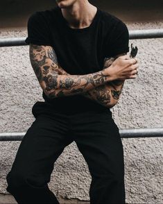 a man with tattoos is leaning against a fence and holding his arms crossed, looking at the camera