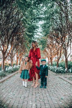 a woman and two children walking down a brick path in front of trees with leaves on the ground