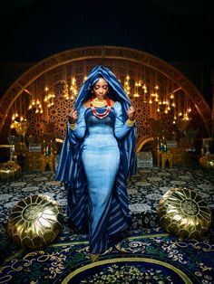 a woman in a blue dress stands on a rug with gold decorations around her and wears an elaborate headdress