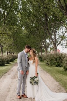 a bride and groom standing in the middle of a dirt road with trees lining both sides