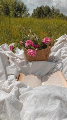 an open book on a blanket with pink flowers in a basket sitting on top of it