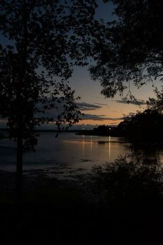 the sun is setting over water with trees in front of it and some lights shining on the water