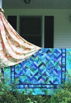 an orange and blue quilt is hanging on the side of a house next to bushes