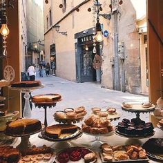 a table full of pastries and desserts on display in an alleyway with people walking by