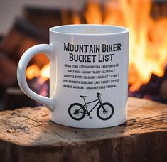 a mountain biker bucket list coffee mug sitting on a log in front of a fire